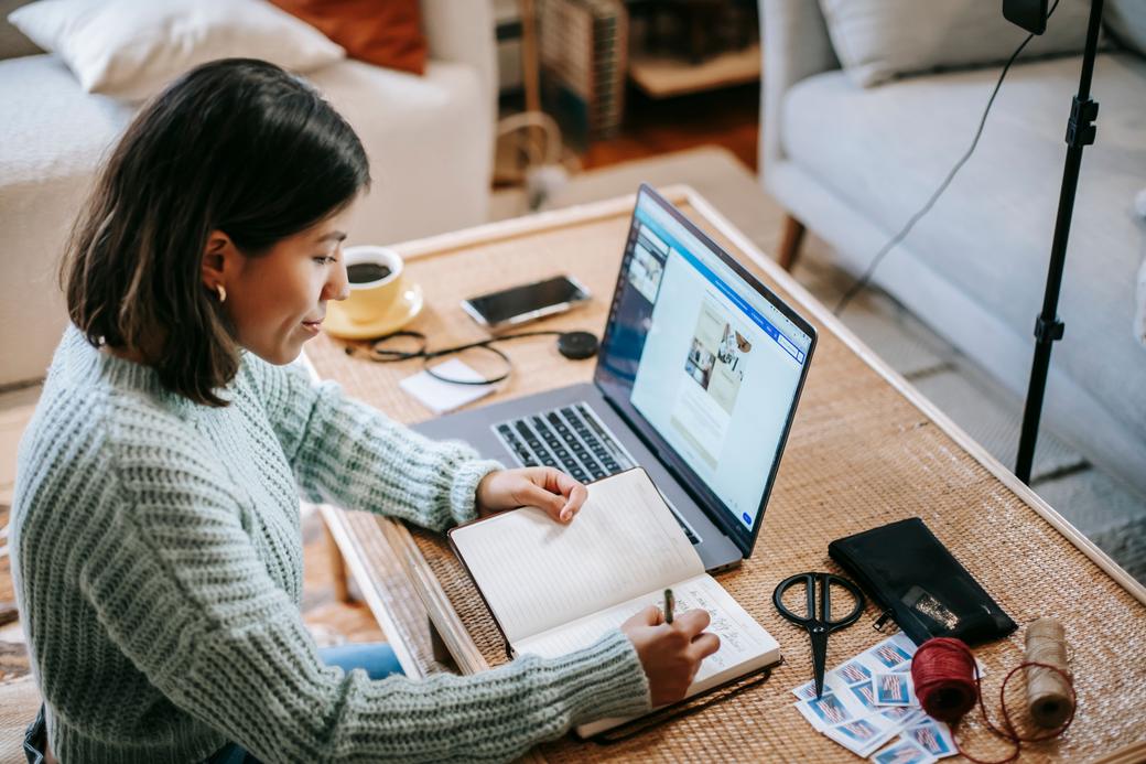 Mujer trabajando en su laptop y tomando notas en una libreta, en el living de su casa